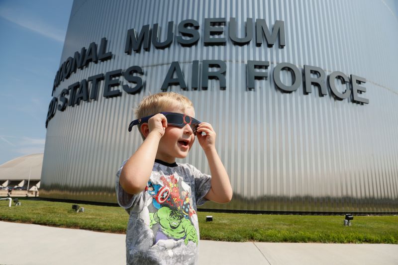 Image of the museum's round silver front with the museum name in black letters and a small boy wearing a Captain America shirt holding up solar ecliplse glasses to his face and looking out into the distance.