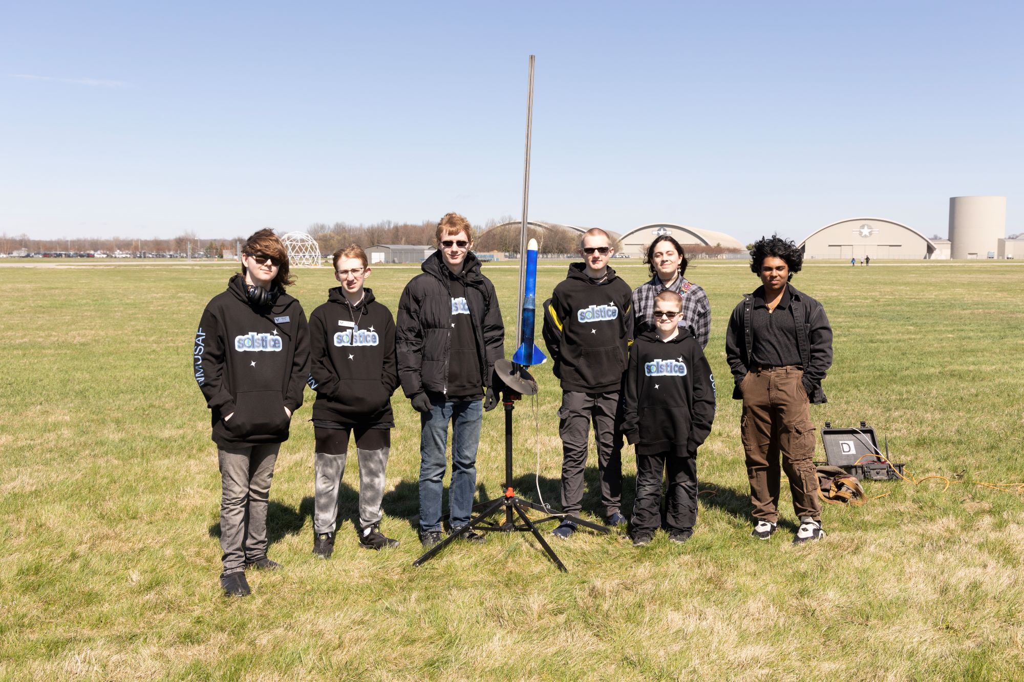 team picture of ARC team Solstice. Seven students standing in front of their blue model rocket with the museum in the background. All are wearing a sweatshirt with the word Solstice on it.