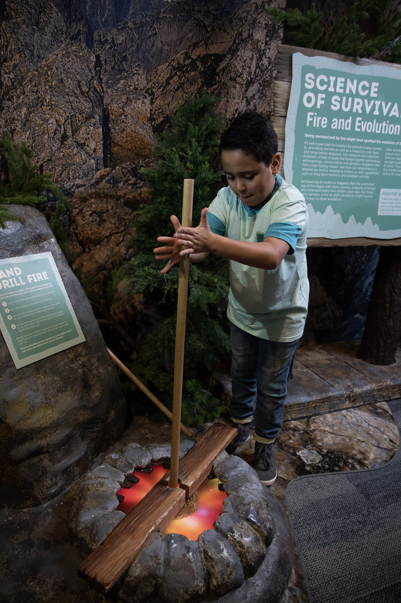 A young boy spins a stick between his hands to practice what it would be like to start a fire  without matches