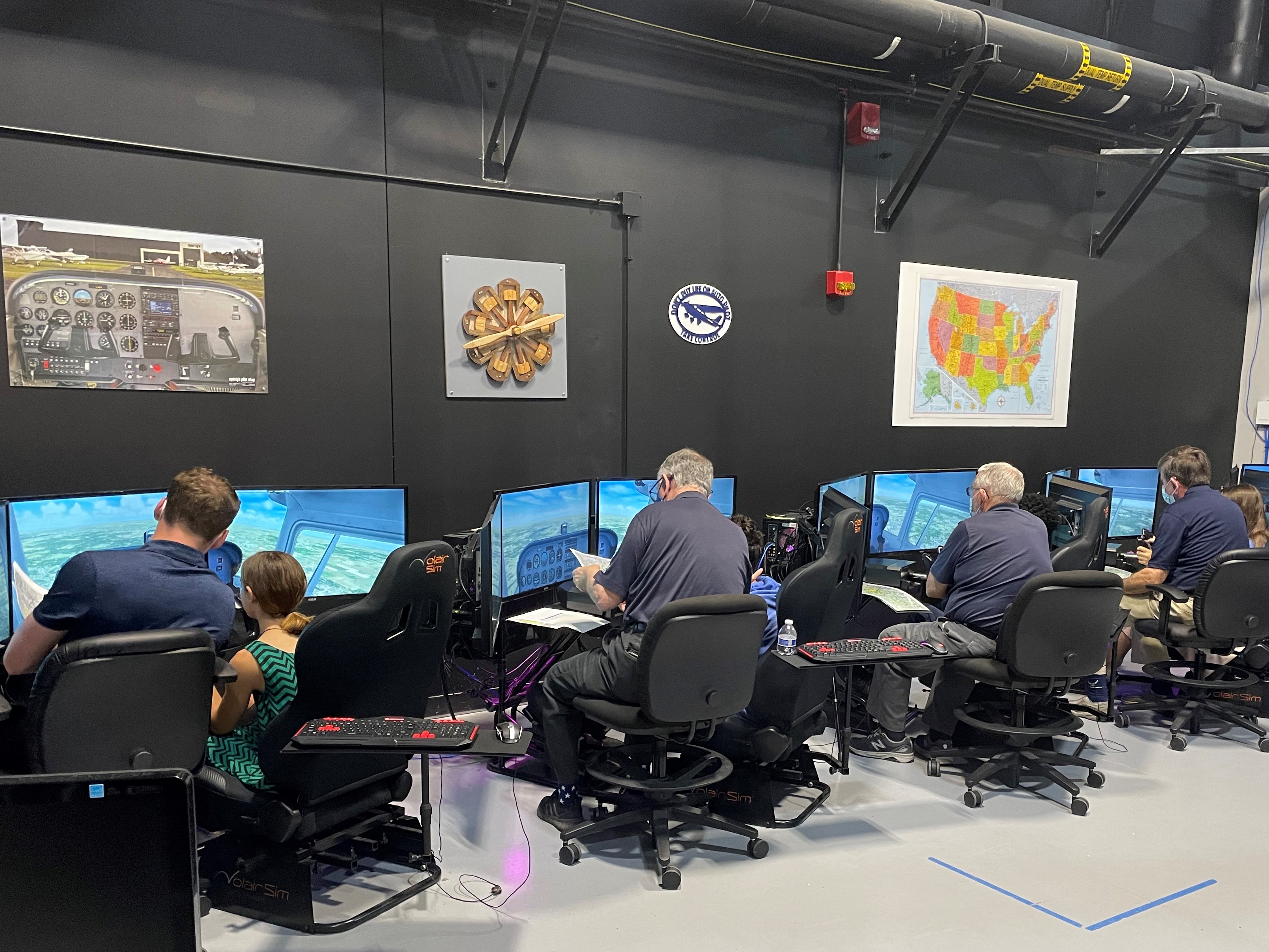 Image of Intro to Pilot course participants and instructors from behind as they look at the monitors showing their flight simulations.