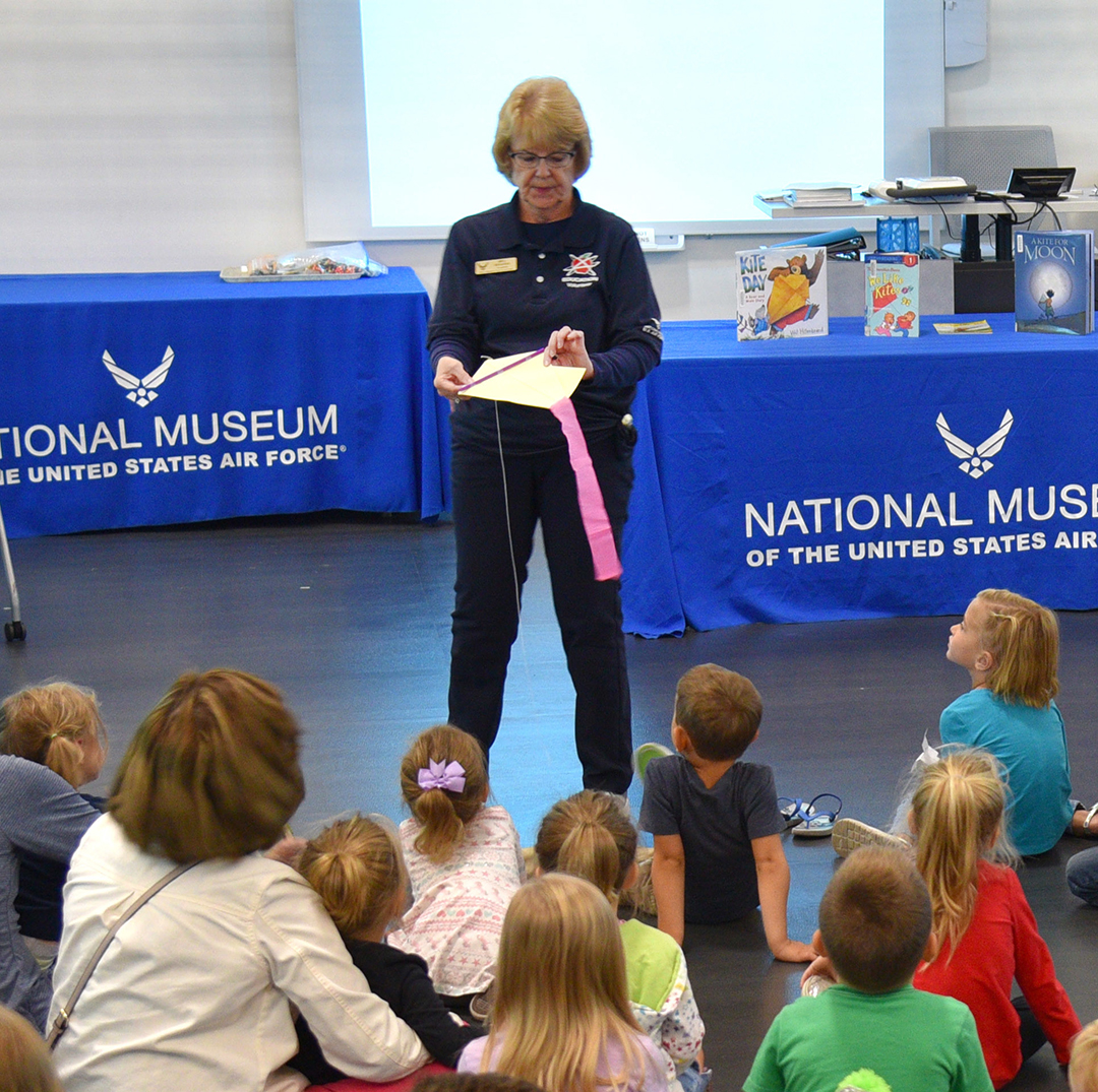 Museum volunteer giving presentation to children sitting on the floor about how to build a kite during storytime in one of the museum's classrooms.