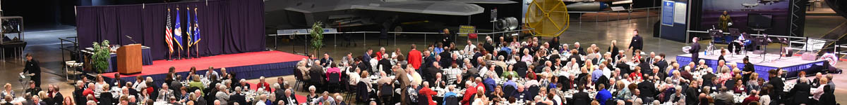 Image of a stage with a podium, black curtain backdrop and US and service flags. There is a large group of people sitting at round banquet tables on the floor in front of the stage.