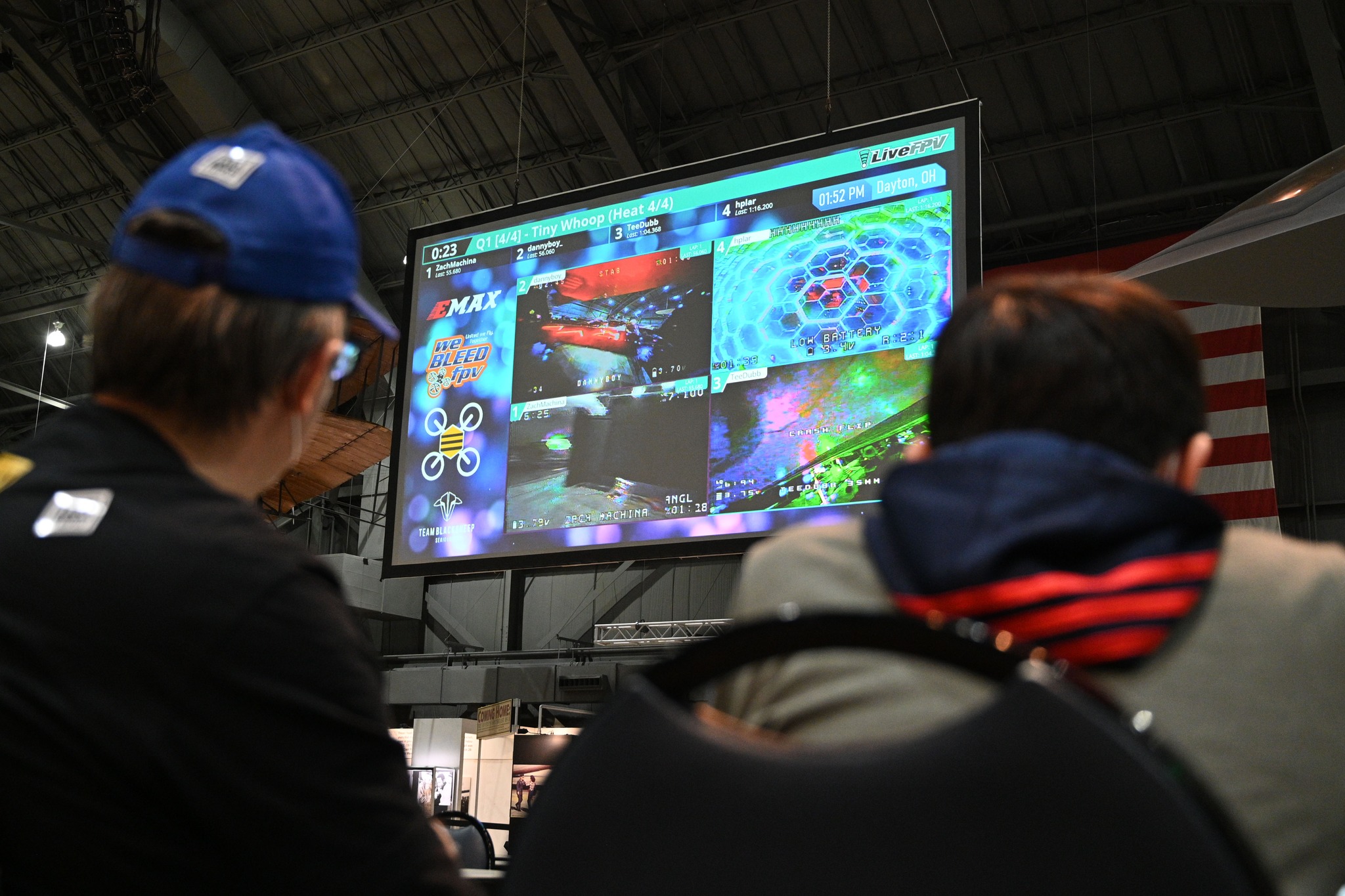 large scrren in background with micro drone races showing on it, in the foreground are people looking up at it.